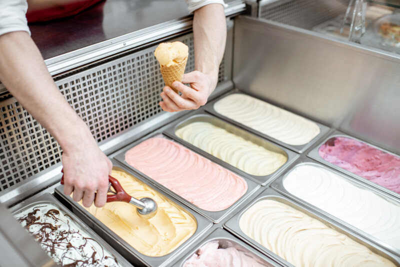 Metal trays full of colorful ice cream in the showcase refrigerator, salesman taking ice cream with scoop, close-up view